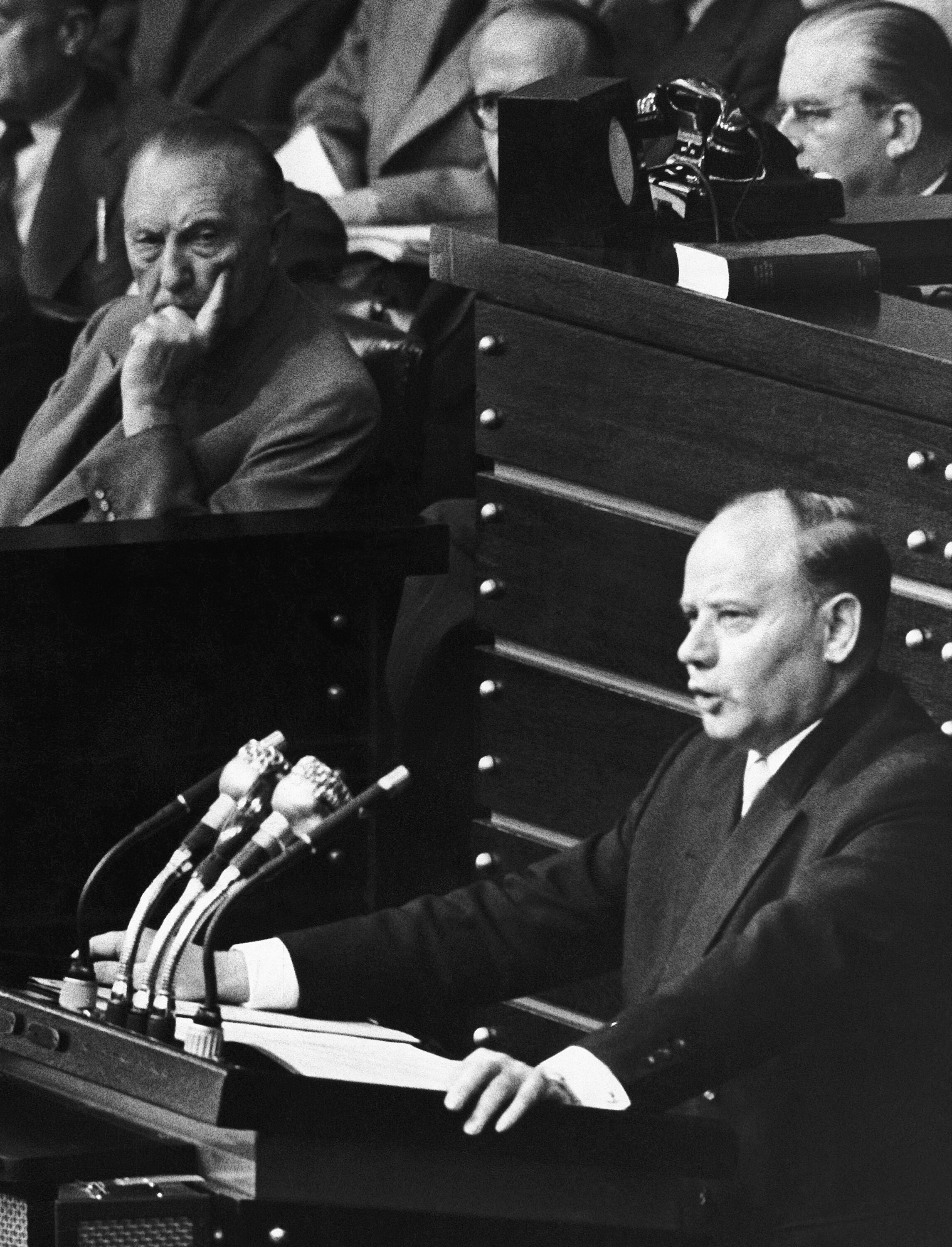 Verteidigungsminister Blank steht an einem Rednerpult, im Hintergrund sitz Bundeskanzler Adenauer.