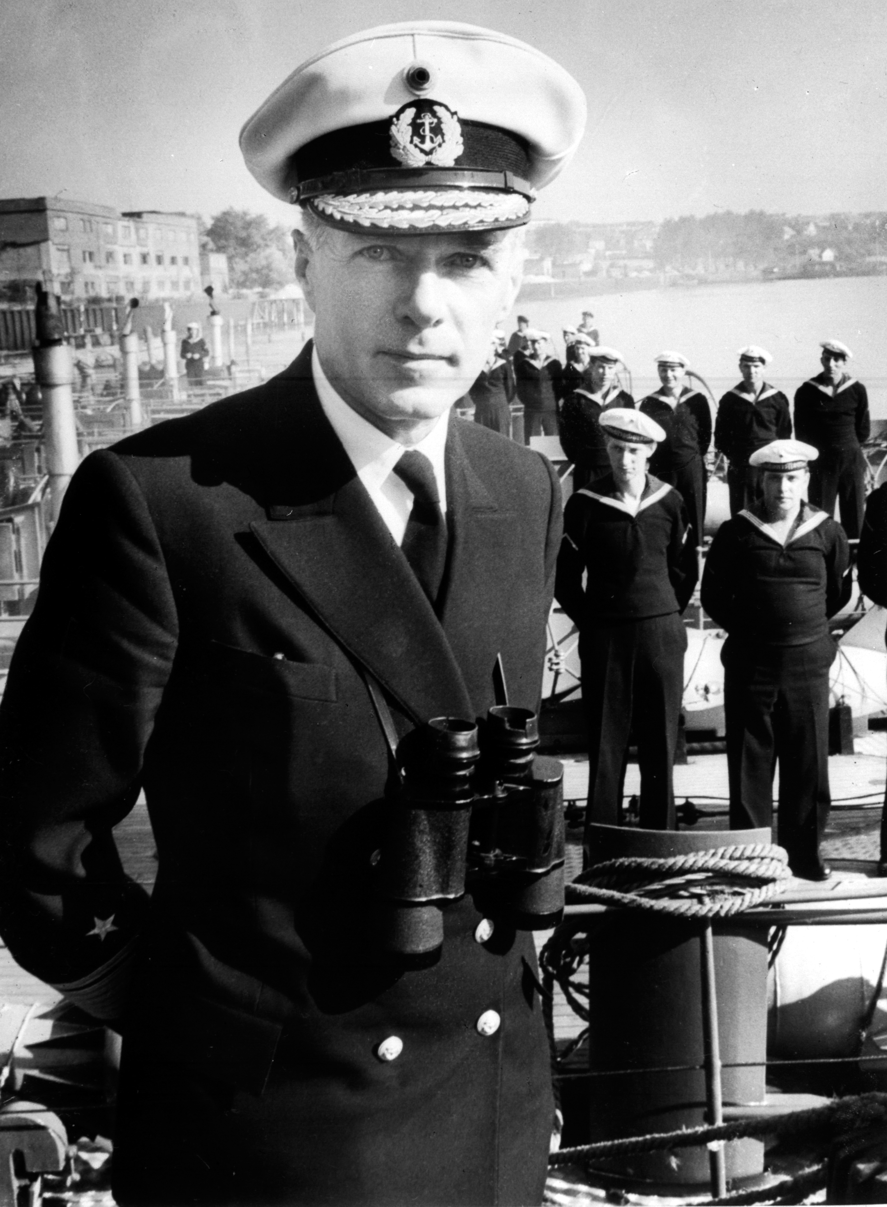Portraitfoto von Admiral Johannesson in Uniform, hinter ihm stehen zahlreiche Matrosen am Kai.