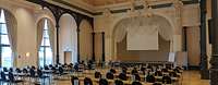 Großer Saal mit verzierten Säulen, Stuck an der Decke und mehreren Stuhlreihen zum Podest gewandt