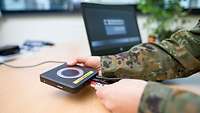 Ein Soldat in Uniform verbindet an einem Schreibtisch ein Laptop mit anderer Hardware.