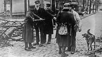 2 Reichswehr Soldaten durchsuchen während des Hamburger Aufstands 4 Passanten und ein Hund in der Nähe einer Barrikade.