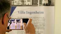 Hinterkopfansicht eines Mannes wie er mit dem Handy Plakate der Villa Ingenheim fotografiert