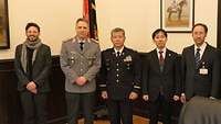 Fünf Männer (zwei in Uniform) und drei Herren im Anzug stehen im Büro vor der Truppenfahne.