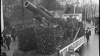 Ein Festumzugswagen auf der Straße mit einer Nachbildung einer schweren Feldhaubitze, die von mehreren Soldaten bedient wird. 