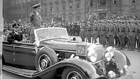 Hitler fährt stehend und den Hitlergruß zeigend an angetretenen Soldaten und Zuschauern vorbei.