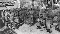 Deutsche Kriegsgefangene beim Marsch durch eine deutsche Ortschaft mit zerstörten Fachwerkhäusern in Richtung einer Brücke