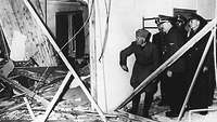 Hitler und Mussolini blicken in die zerstörten Überreste des Konferenzraums in der Wolfsschanze, der von Trümmern übersät ist.