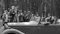 Dreharbeiten zum Film, zu sehen sind Statisten. Ein Kameramann hält eine Filmkamera auf den Darsteller von Stauffenberg im Auto.