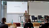 Mann mit Brille steht vor einer Powerpoint Präsentation am Podium im Vordergrund ist eine Rückansicht des Publikums zu sehen.