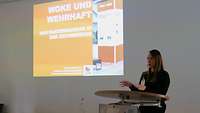 Eine Frau steht am Rednerpult und trägt mit Unterstützung einer Präsentation am Beamer vor.