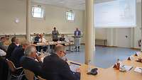 Ein kleiner Saal, in dem mehrere Personen an Tischen sitzen und einem Vortrag zuhören.