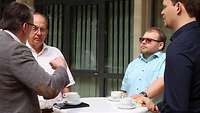 Vier Personen stehen im Kreis. Eine Person im Bild spricht und gestikuliert, die anderen hören zu