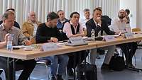 Fünf Personen sitzen nebeneinander an einem Tisch, hören sich einen Vortrag an und machen Notizen