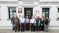 12 Personen, gemischt in Uniform und zivil, posieren für ein Gruppenfoto vor einem alten Haus.