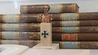 In einer Vitrine liegen 11 alte Bücher übereinander hinter einer Miniaturbüste von Clausewitz