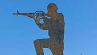 Silhouette eines Soldaten, der eine Waffe im Anschlag hält aus Metall auf einem Steinhügel.
