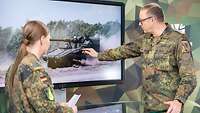 Eine Soldatin interviewt einen Soldaten, der an einem Monitor etwas an einem Panzer zeigt