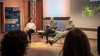Drei Personen sitzen auf einem Podium, darunter eine Frau und ein Offizier in Uniform