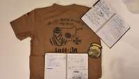 Handschriftliche Aufzeichnungen und ein Klett-Patch liegen auf einem bedruckten, ockerfarbenen Shirt