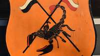 Ein schwarzer Skorpion vor gekreuzten Fahnenlanzen auf orange-gelbem Holzschild
