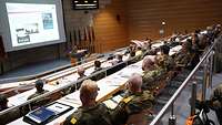 Etwa 50 Menschen folgen in einem Hörsaal einer Präsentation durch einen Soldaten am Rednerpult.