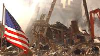 Trümmer des World Trade Centers ragen in die Luft, davor arbeiten Helfer, links weht die US-Flagge.