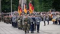 Viele Männer in grünen und grauen Uniformen marschieren in einer Reihe, im Hintergrund Flaggen