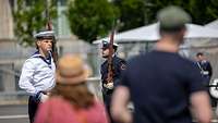 Eine Frau und ein Mann betrachten zwei Soldaten des Wachbataillons in Marineuniform mit Gewehr