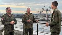 Drei Soldaten stehen zusammen und sprechen miteinander. Im Hintergrund ist ein Hafen zu sehen.